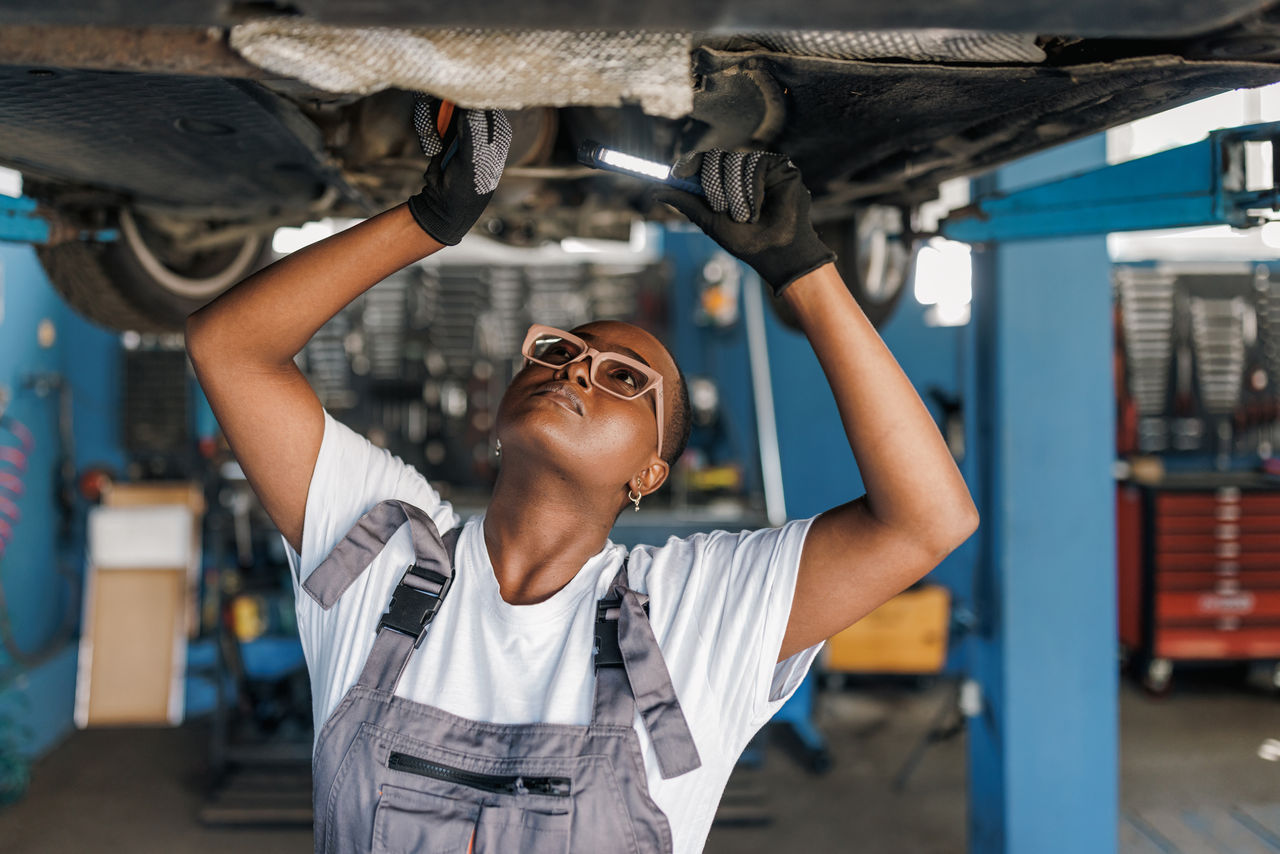 false A young African American female mechanic is standing under a car and examining the damage on the broken parts with a serious look on her face.