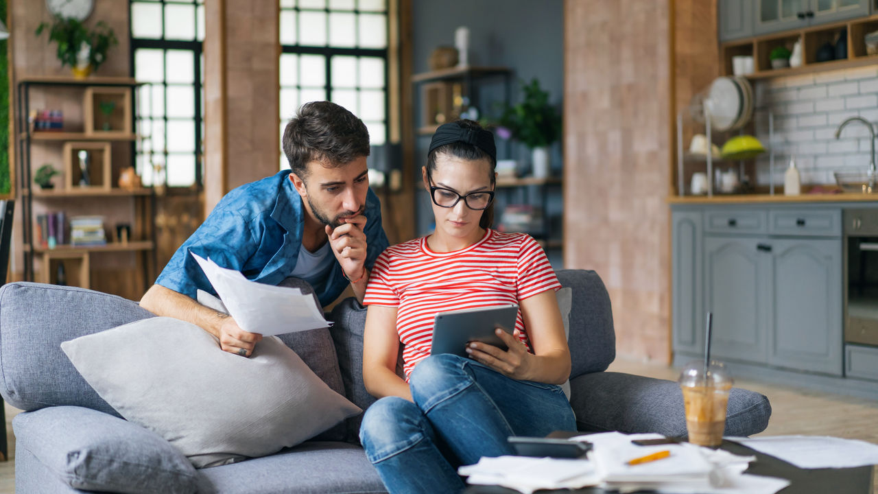 false Couple calculating bills at home using laptop and calculator. Young couple working on computer while calculating finances sitting on couch. Young man with wife at home analyzing their finance with documents.