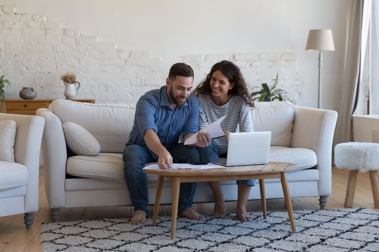 false Millennial wife and husband sorting out bank papers, check invoices, paying bills via e-bank on laptop sit on sofa smile feel satisfied. Enough money, payments, profit, financial management concept
