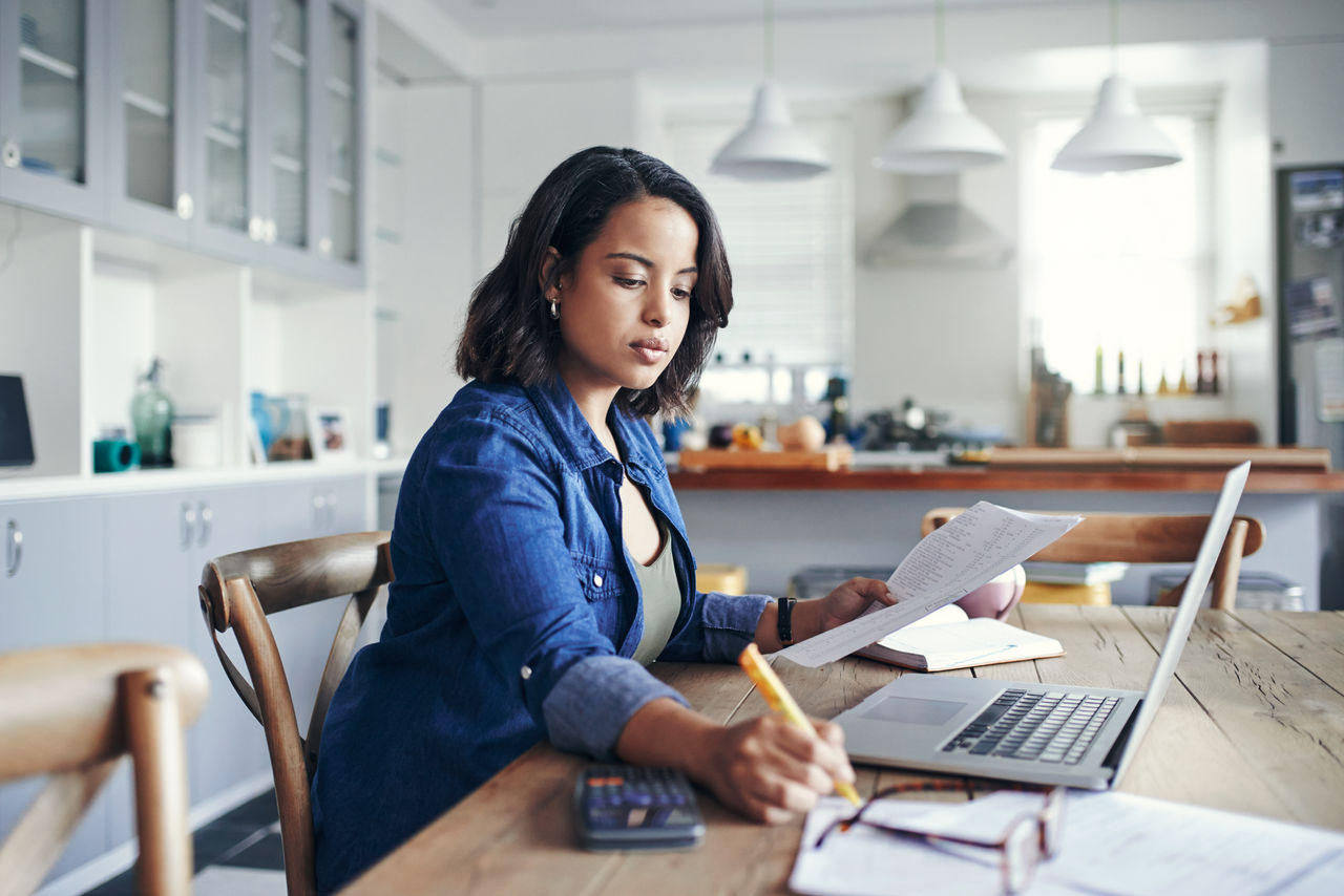 false Shot of a young woman using a laptop and going through paperwork while working from home