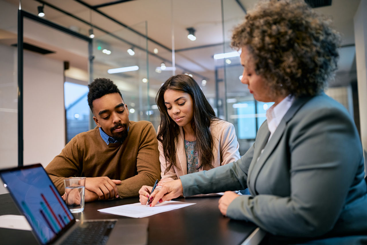1483243964 African American woman and her husband signing mortgage agreement during a meeting with their bank manager.