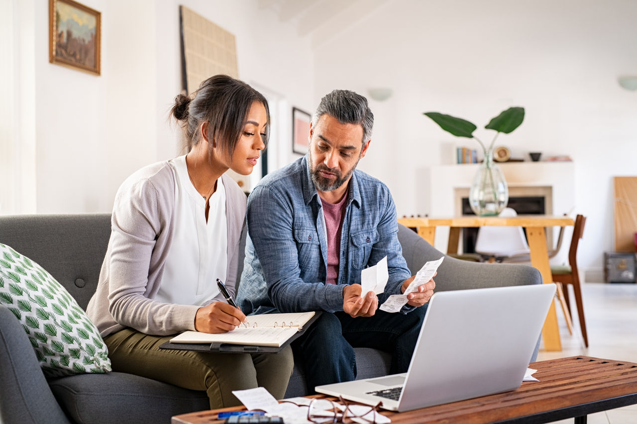 false Mature couple calculating bills at home using laptop and calculator. Multiethnic couple working on computer while calculating finances sitting on couch. Mature indian man with african american woman at home analyzing their finance with documents.