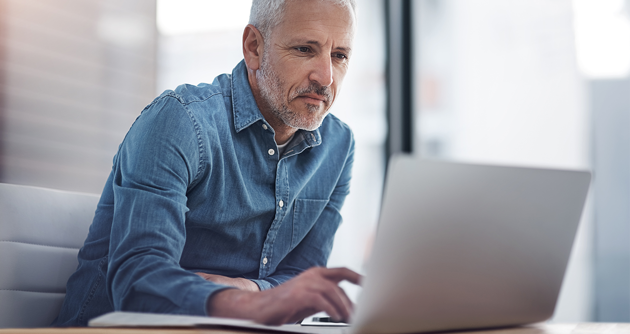 gray-haired man in blue denim shirt looking at silver-colored laptop computer